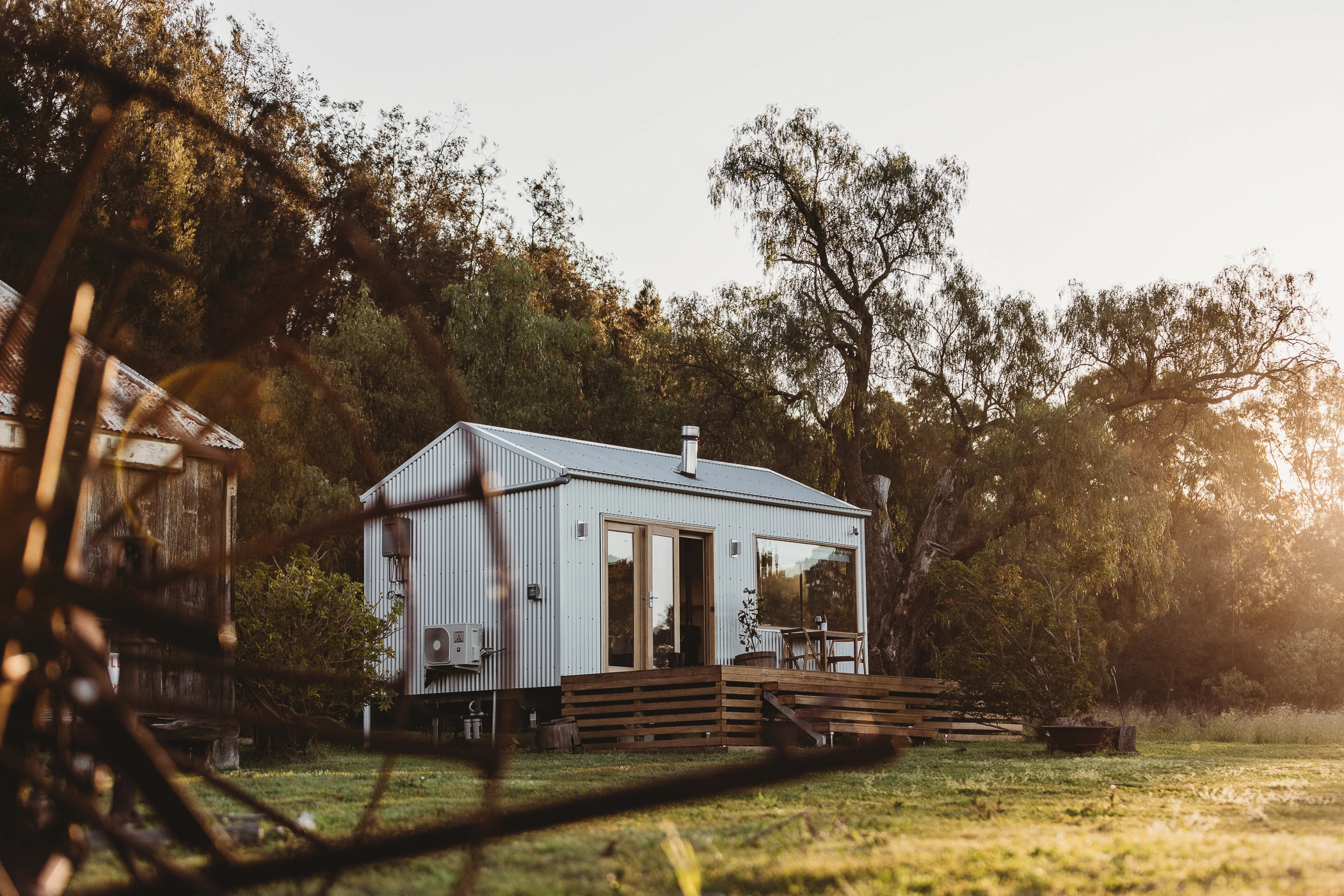 White cabin with a timber deck among trees in late afternoon sunlight
